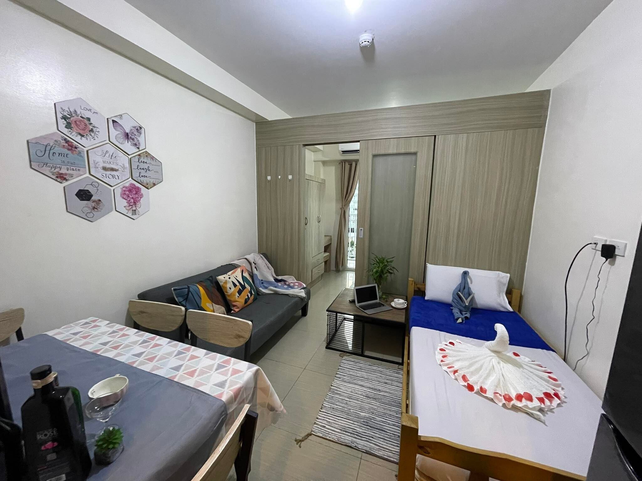 A modern living room with a sofa, a dining table, and a bed. There are hexagonal wall decorations and a laptop on the table.