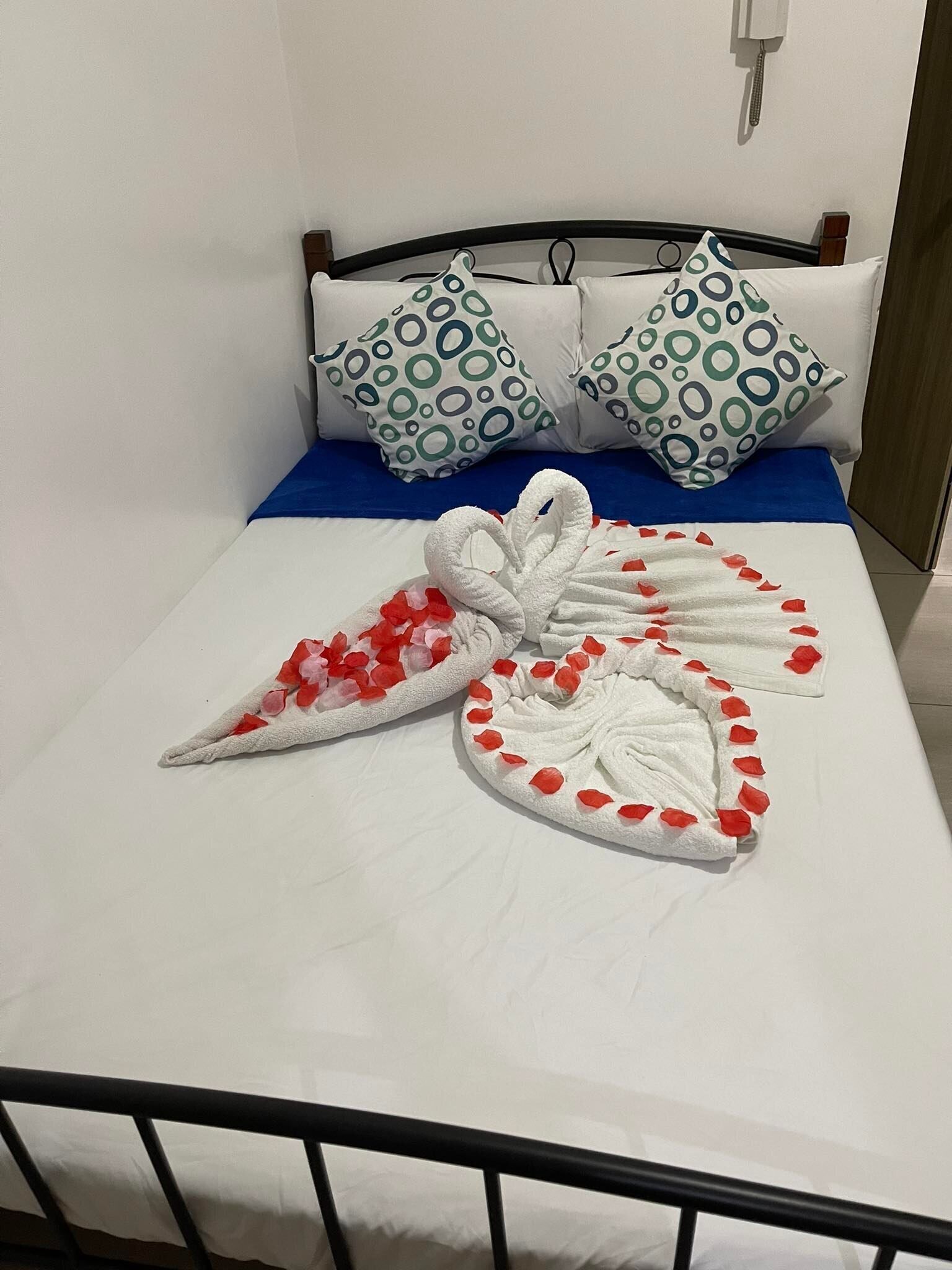 A neatly made bed with a blue bedspread, two pillows with green and white circular patterns, and two towels arranged in a heart shape with red rose petals.