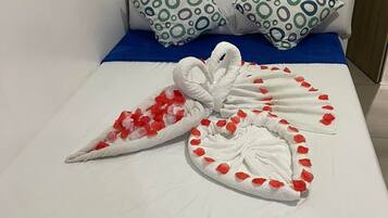 A neatly made bed with a blue bedspread, two pillows with green and white circular patterns, and two towels arranged in a heart shape with red rose petals.