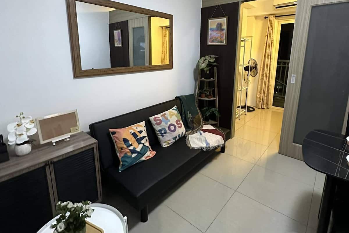 Una sala moderna con un sofá negro, una mesa de centro blanca con una planta, un espejo grande y una puerta que da a otra habitación.