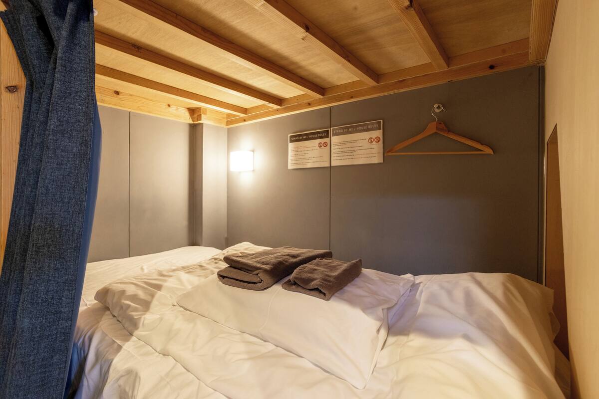 Uma cama bem arrumada com lençóis brancos e duas toalhas dobradas, vigas de madeira no teto e uma placa na parede com as regras da hospedagem.
