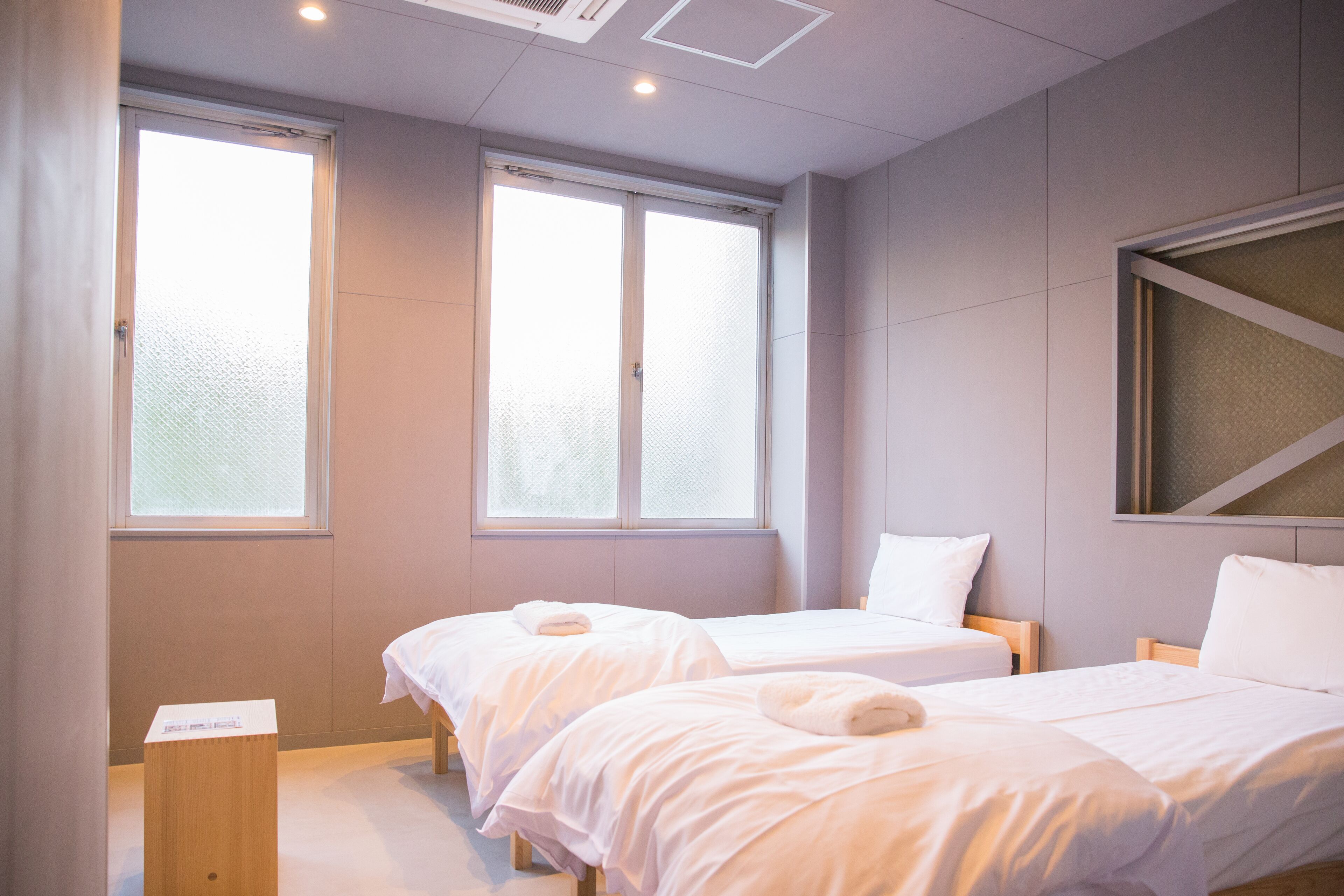 A hotel room with two beds, a wooden nightstand, and a window with frosted glass.