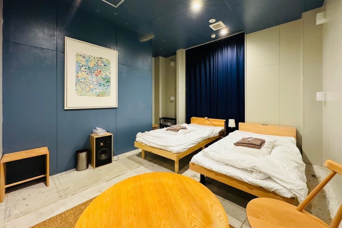 Quarto moderno com duas camas, piso de madeira, pintura abstrata emoldurada, um pequeno alto-falante e uma mesa de madeira redonda.