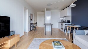 Un appartement moderne comprenant un salon, un coin repas et une cuisine. On y trouve une télévision, une bibliothèque et une table à manger avec des chaises.