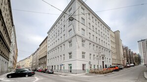 Un grand immeuble à étages, de style architectural classique, qui se distingue par son entrée d’angle imposante et une horloge sur sa façade.