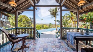 A covered outdoor area with a wooden bench, a table, and a view of a swimming pool and palm trees. - BICKERTON ESTATE (Pecatu)