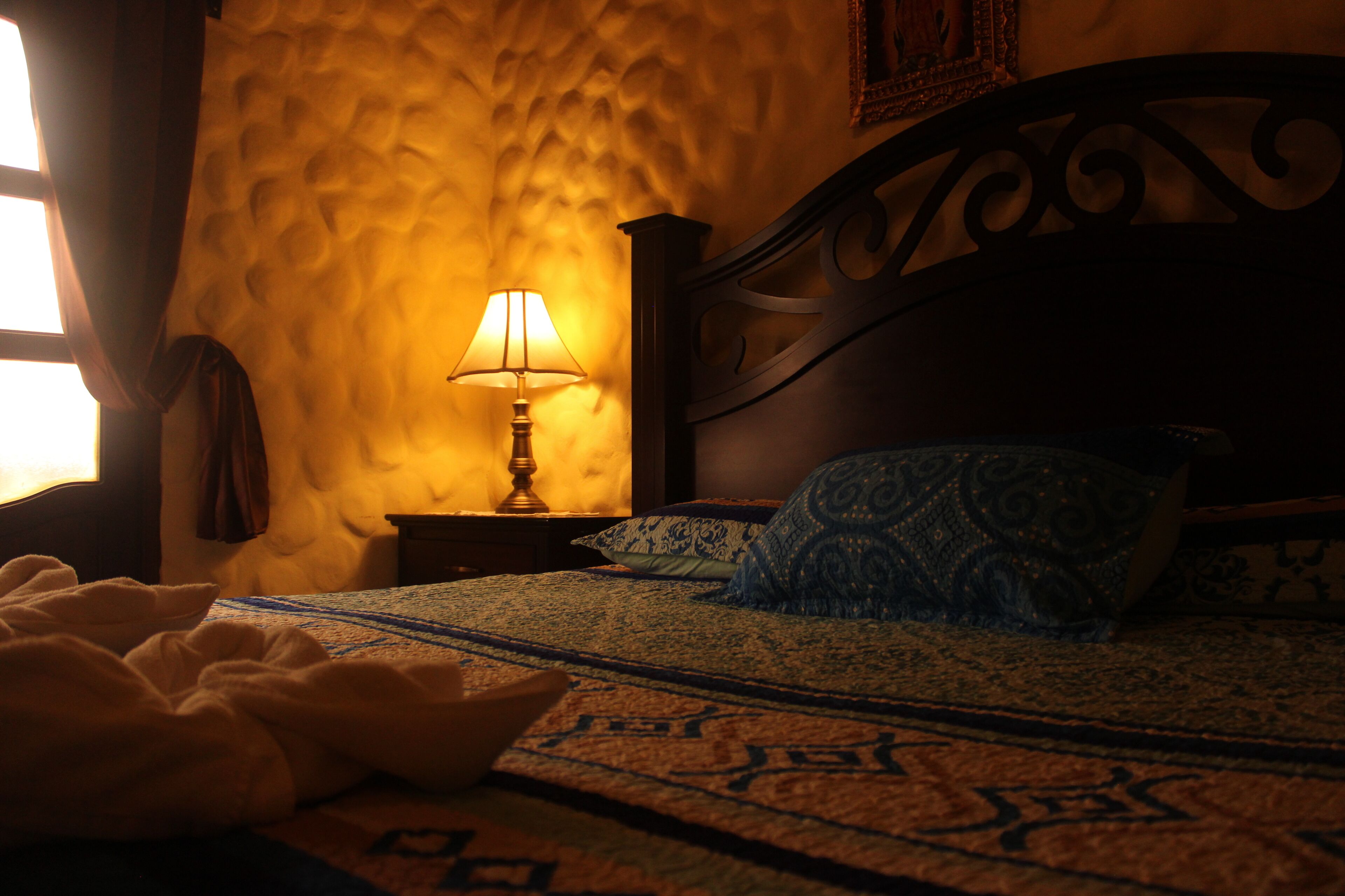 Un dormitorio con una colcha estampada, una lámpara de noche y una ventana con cortinas.