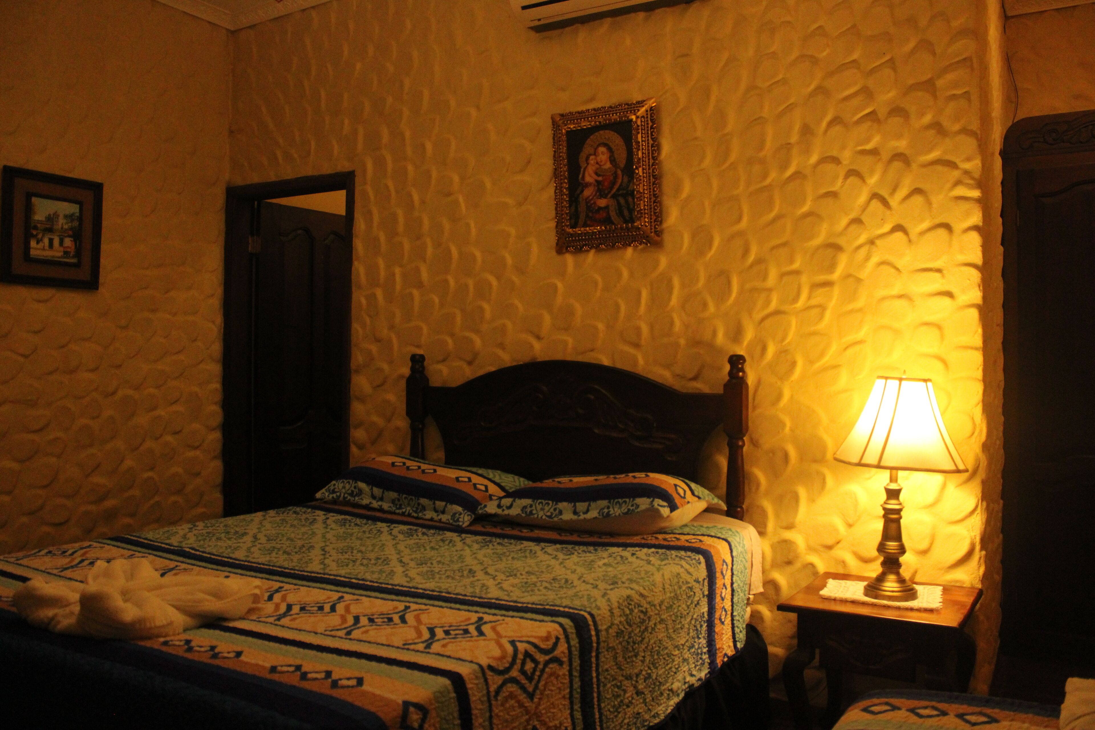 Una habitación con pared texturizada, una cama con ropa de cama estampada, una mesita de noche con lámpara y un cuadro enmarcado sobre la cama.