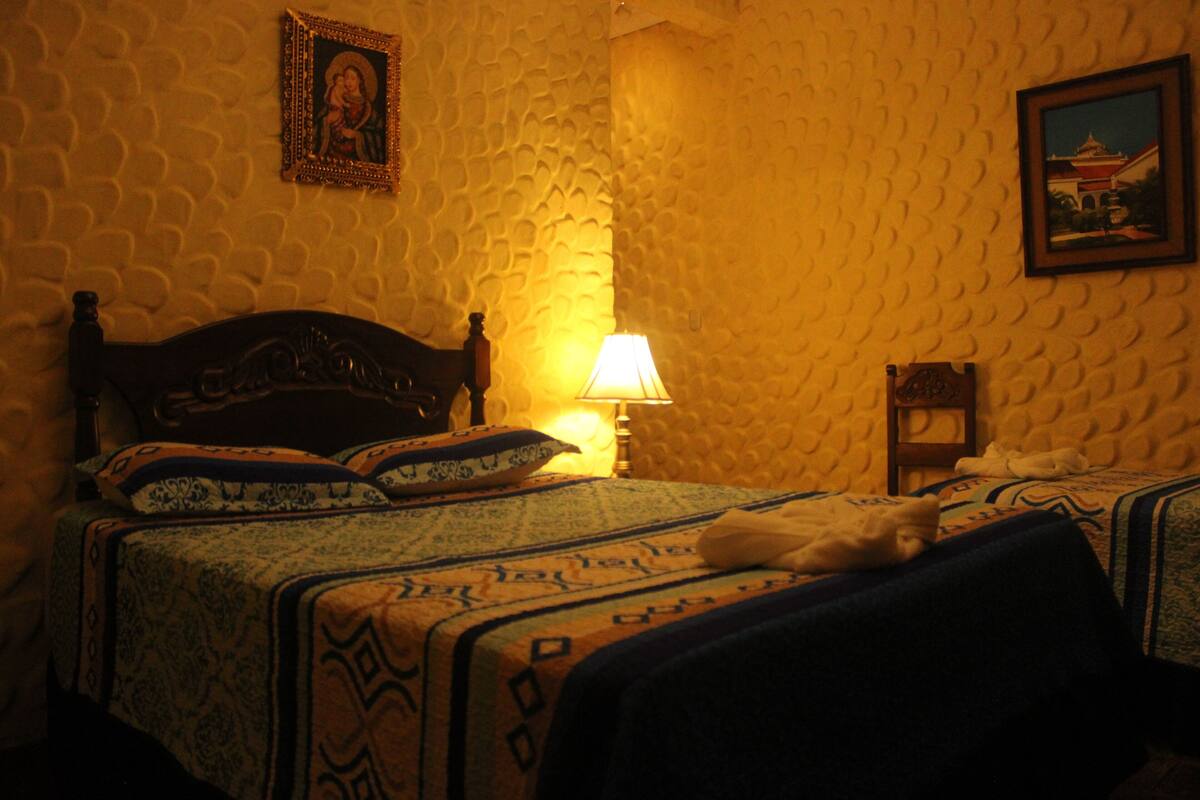 A room with a patterned wall, a bed with a patterned bedspread, a bedside lamp, and a framed picture on the wall.