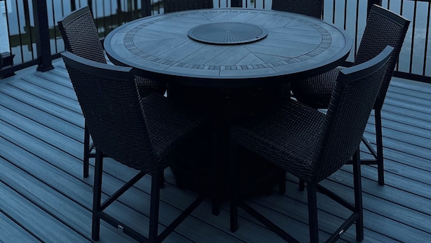 Une table ronde entourée de chaises, installée sur une terrasse en bois qui donne sur un lac paisible, avec un quai visible au loin.