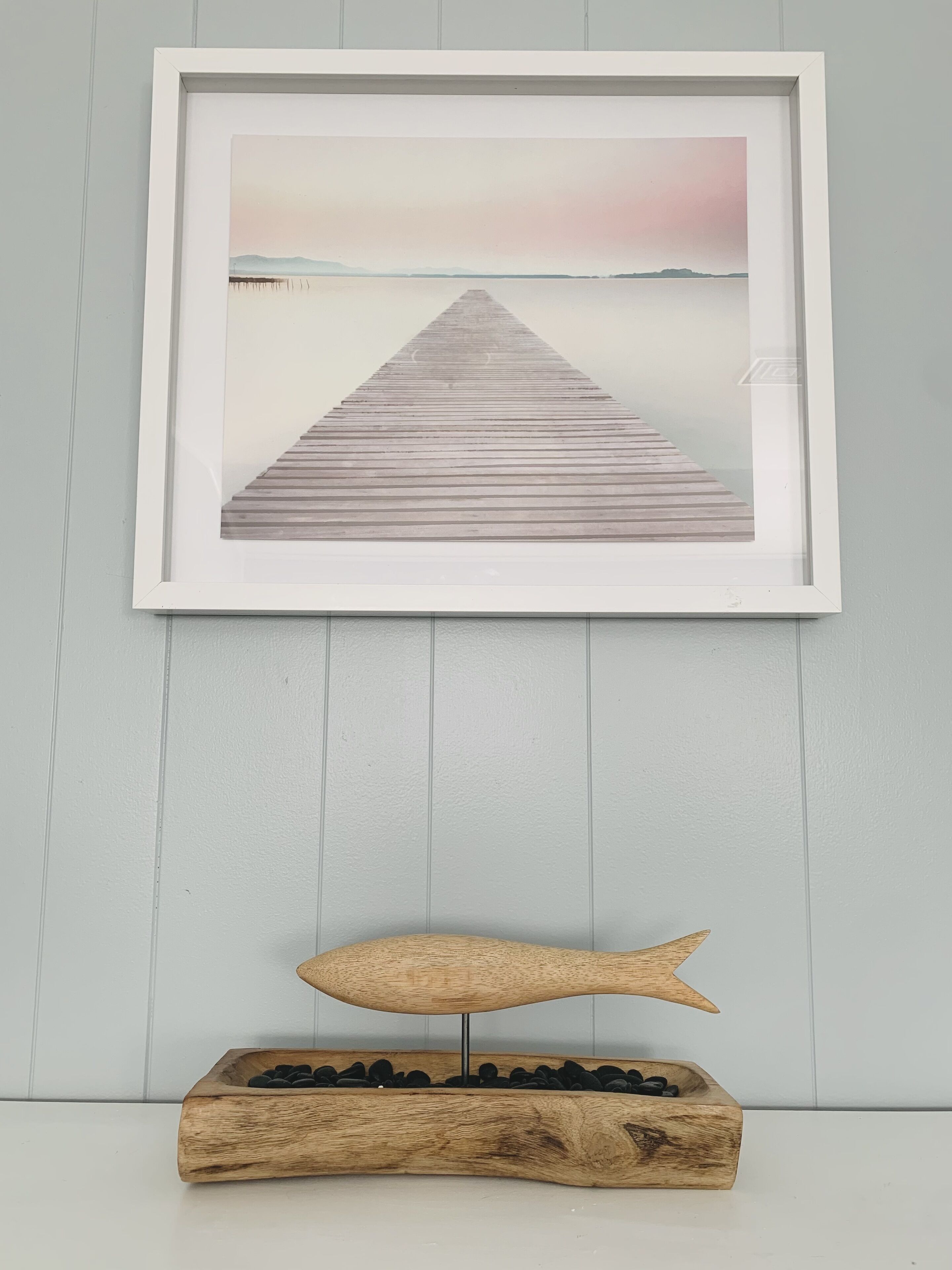 Een lijst met een foto van een houten steiger die zich uitstrekt over kalm water, met een houten sculptuur van een vis eronder.