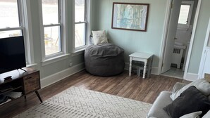 Un salon spacieux avec un grand canapé gris, un fauteuil en forme de poire et un téléviseur posé sur un meuble en bois. De grandes fenêtres offrent une vue sur l’extérieur.