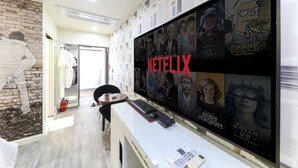 Huoneessa on suuri seinälle asennettu televisio, jossa näkyy erilaisia Netflixin ohjelmia, työpöytä, jossa on tietokone, ja lattialla palosammutin.