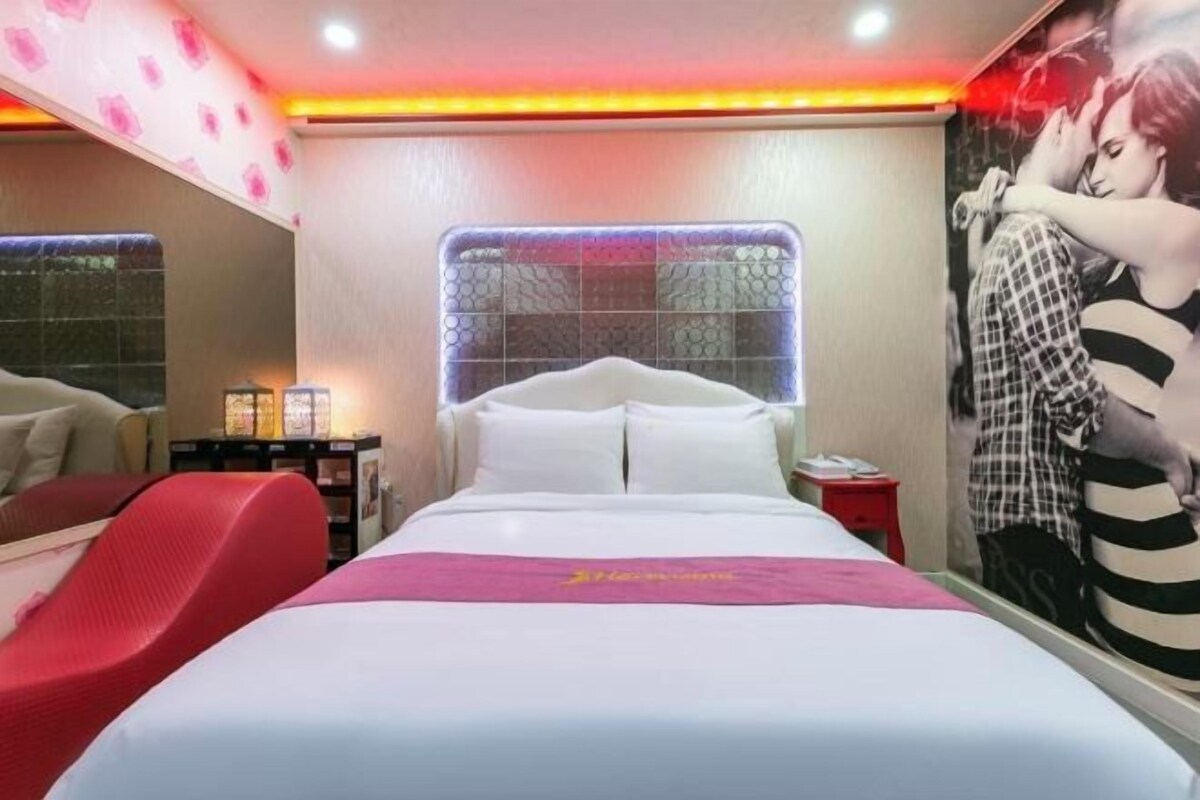 A hotel room with a large bed, a red chair, a nightstand, and a wall-mounted mirror.