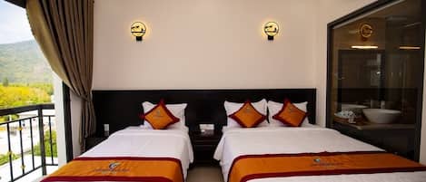 A hotel room with two beds, each with white bedding and orange accents, a balcony with a view of greenery, and wall-mounted lighting.