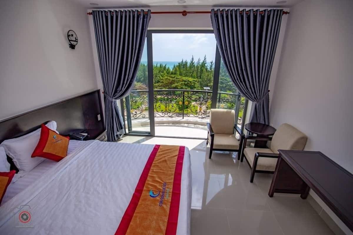 Ein Hotelzimmer mit Bett, Stuhl, kleinem Tisch und einem Balkon mit Vorhängen.