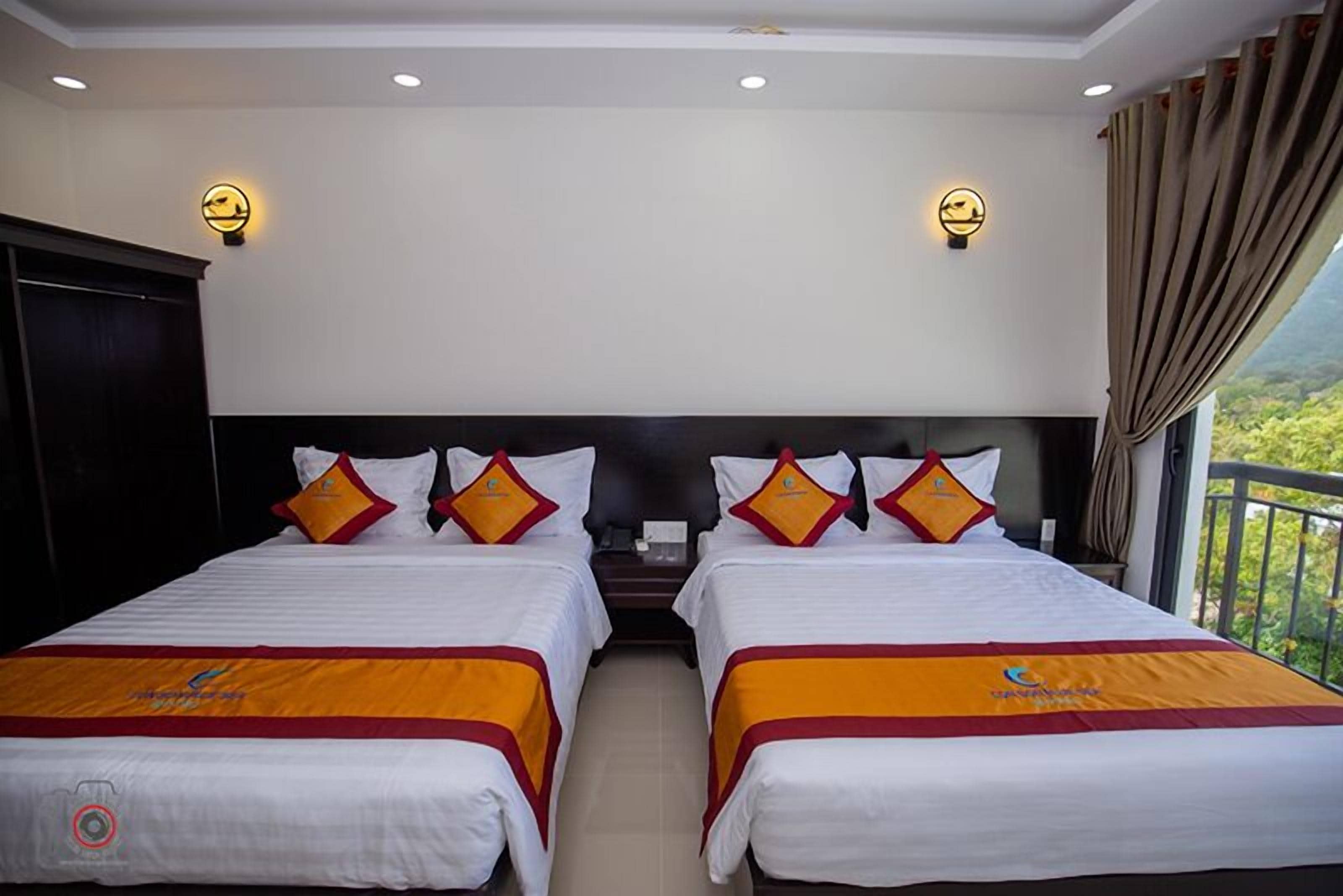A hotel room with two beds, each with a headboard, white bedding, and orange bedspreads. There are wall-mounted light fixtures and a view of greenery outside.