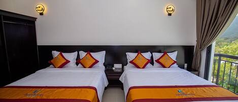 A hotel room with two beds, each with a headboard, white bedding, and orange bedspreads. There are wall-mounted light fixtures and a view of greenery outside.