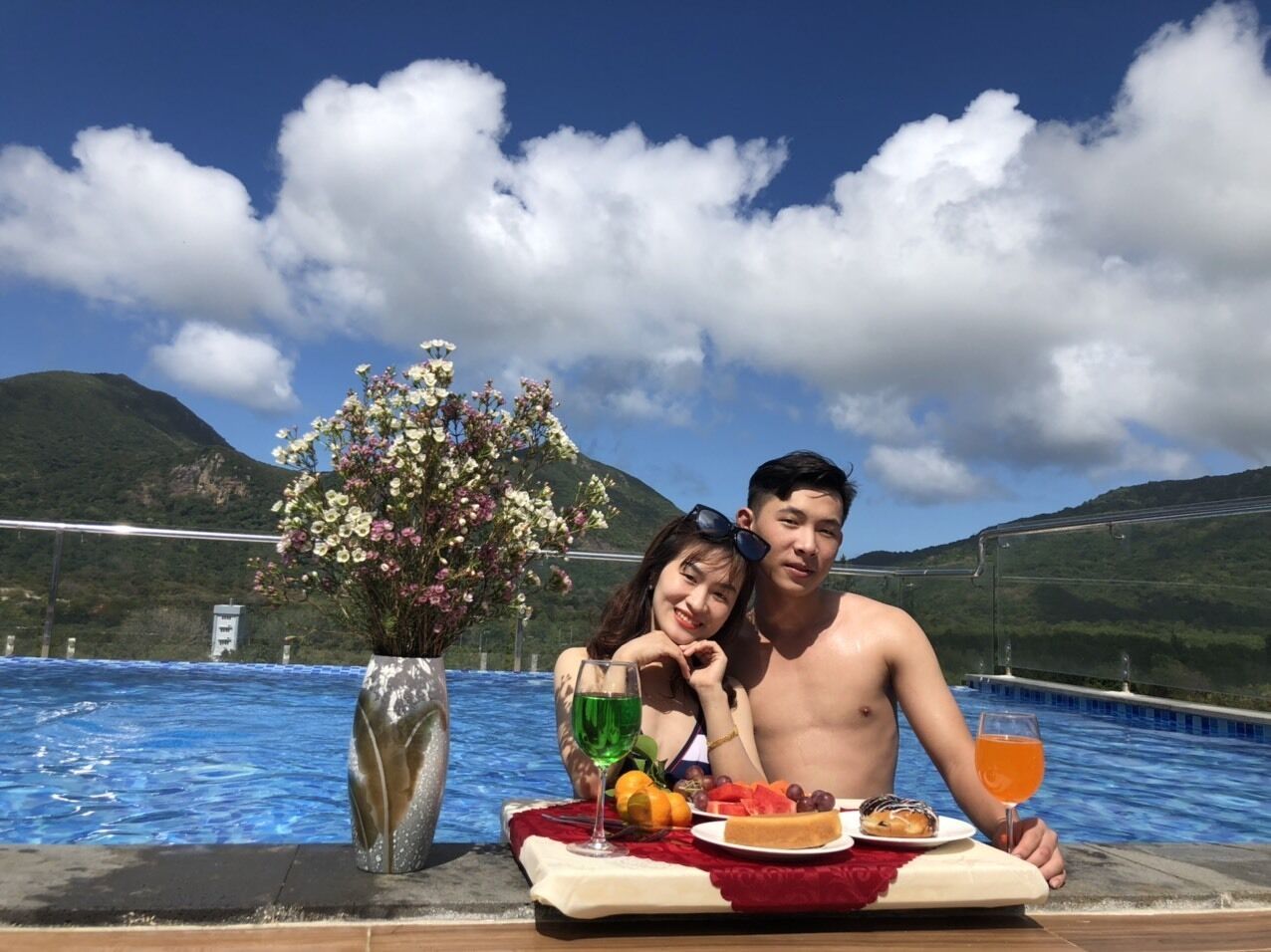 Un couple est assis à une table au bord de la piscine, devant des boissons, des fruits et un vase rempli de fleurs.