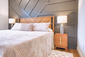 A neatly made bed with a wooden headboard, two bedside lamps, and a checkered rug. - Modern Wooded Retreat | Corporate Housing | Fenced Yard, Pets OK | Near Back 40 (Bella Vista)