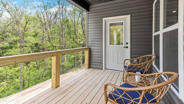 A wooden deck with a railing, two wicker chairs with cushions, and a door leading to a covered area with a view of trees.