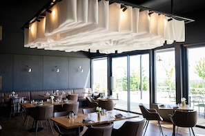 An indoor dining area with tables and chairs, large windows, and a ceiling with hanging fabric.