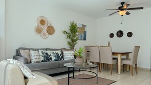 A living room with a grey sofa, a coffee table, a dining table with chairs, a ceiling fan, and wall art. - Alojamiento con Vistas Costa del Este 1 (Panamá)