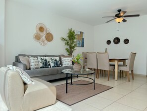 Un salon avec un canapé gris, une table basse, une table à manger avec des chaises, un ventilateur de plafond et des œuvres d’art murales.