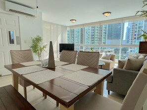 A modern living room with a dining area, large windows, and a view of high-rise buildings.