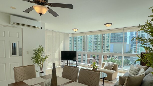 A modern living room with a dining area, large windows, a ceiling fan, and a flat-screen TV. - Alojamiento con Vistas Costa del Este 1 (Panamá)