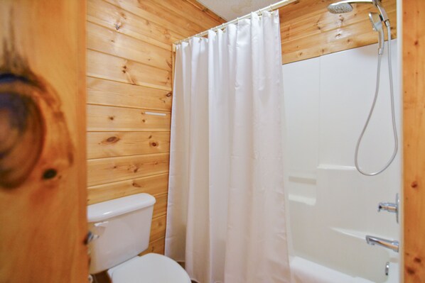 Une salle de bain aux murs lambrissés, équipée de toilettes, d’une douche avec rideau et d’une baignoire.