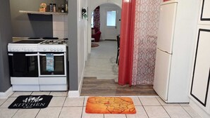 A kitchen with a gas stove, oven, and refrigerator. There is a red curtain and a patterned curtain in the doorway. A map rug is on the floor.