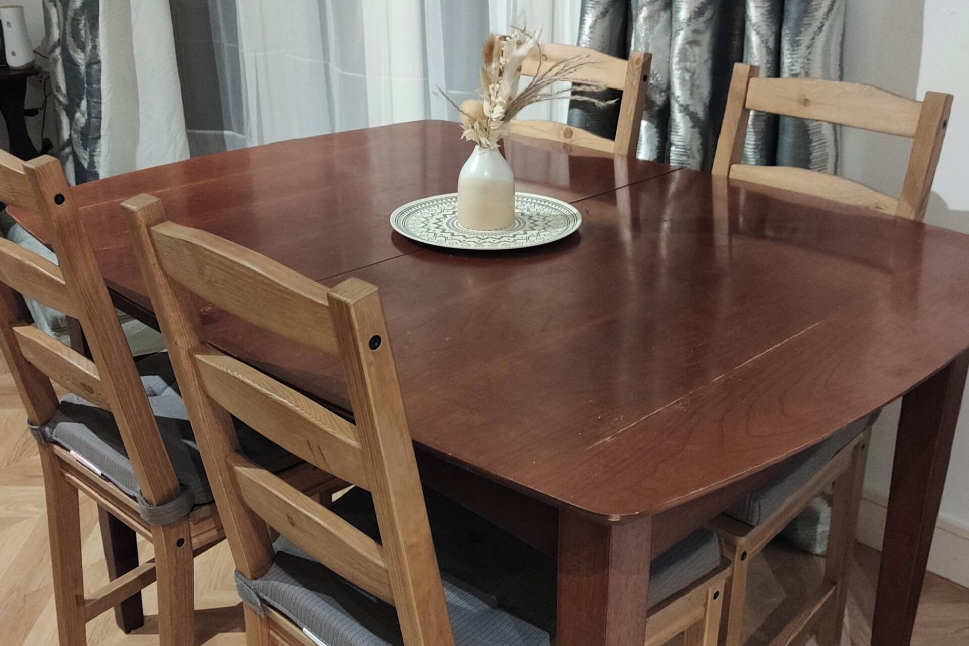 A wooden dining table with a white vase and dried flowers on a patterned coaster.