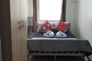 A small, neatly made bed with a grey bedspread and multiple patterned pillows. There is a bedside table with a lamp and a power outlet. The room has a simple, functional design with a wooden wardrobe and a window with curtains. - Apartment - Sleeps 4 - Parking - Station Close (London)