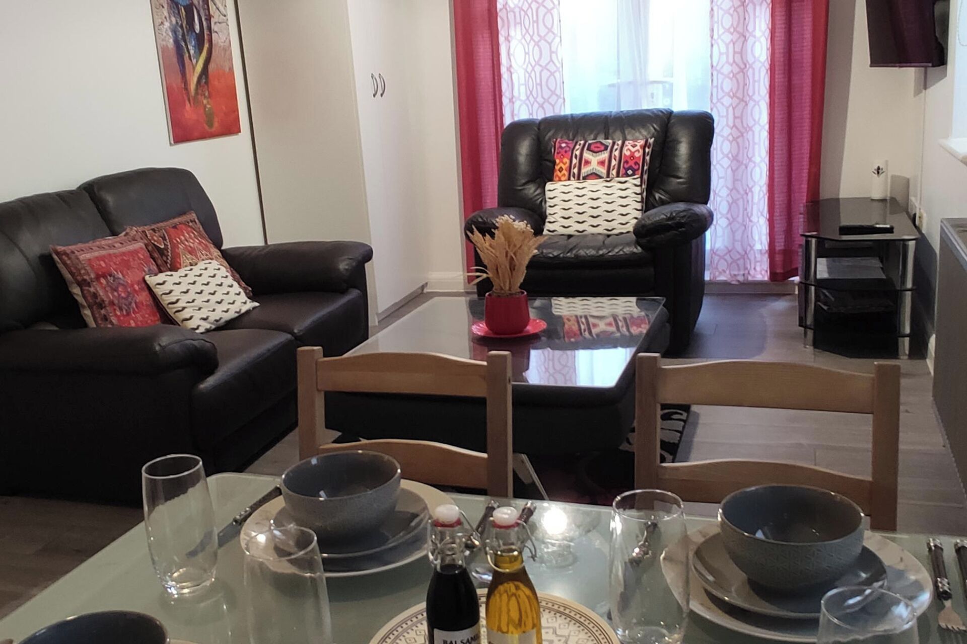 A dining area with a glass table set for two, two black bowls, two wine bottles, and a vase with dried plants.