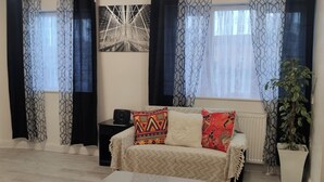 A living room with a sofa, a coffee table, and a radiator. There are curtains on the windows, and a plant is visible.
