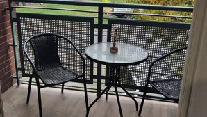 A balcony with two black metal chairs and a small round table, a vase with dried flowers, and a view of a fenced yard and trees.