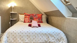 Et soveværelse med skrå væg, seng med mønstret sengetøj, to puder med et rødt og sort design, og en sengetavlelampe.