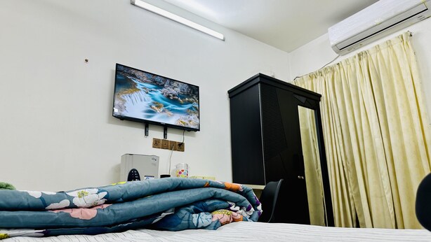 A hotel room with a bed, a TV mounted on the wall, a wardrobe, and a window with curtains. - Beautiful 1-Bed Studio in Dhaka (Dhaka)