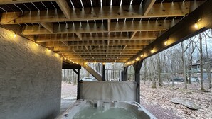 A hot tub under a wooden pergola with string lights, surrounded by trees and a stone wall.