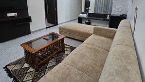 A modern living room with a sofa, a glass-top coffee table, a black entertainment unit, and a mirror reflecting a bathroom. - Annapurna Executive (Tuljapur)