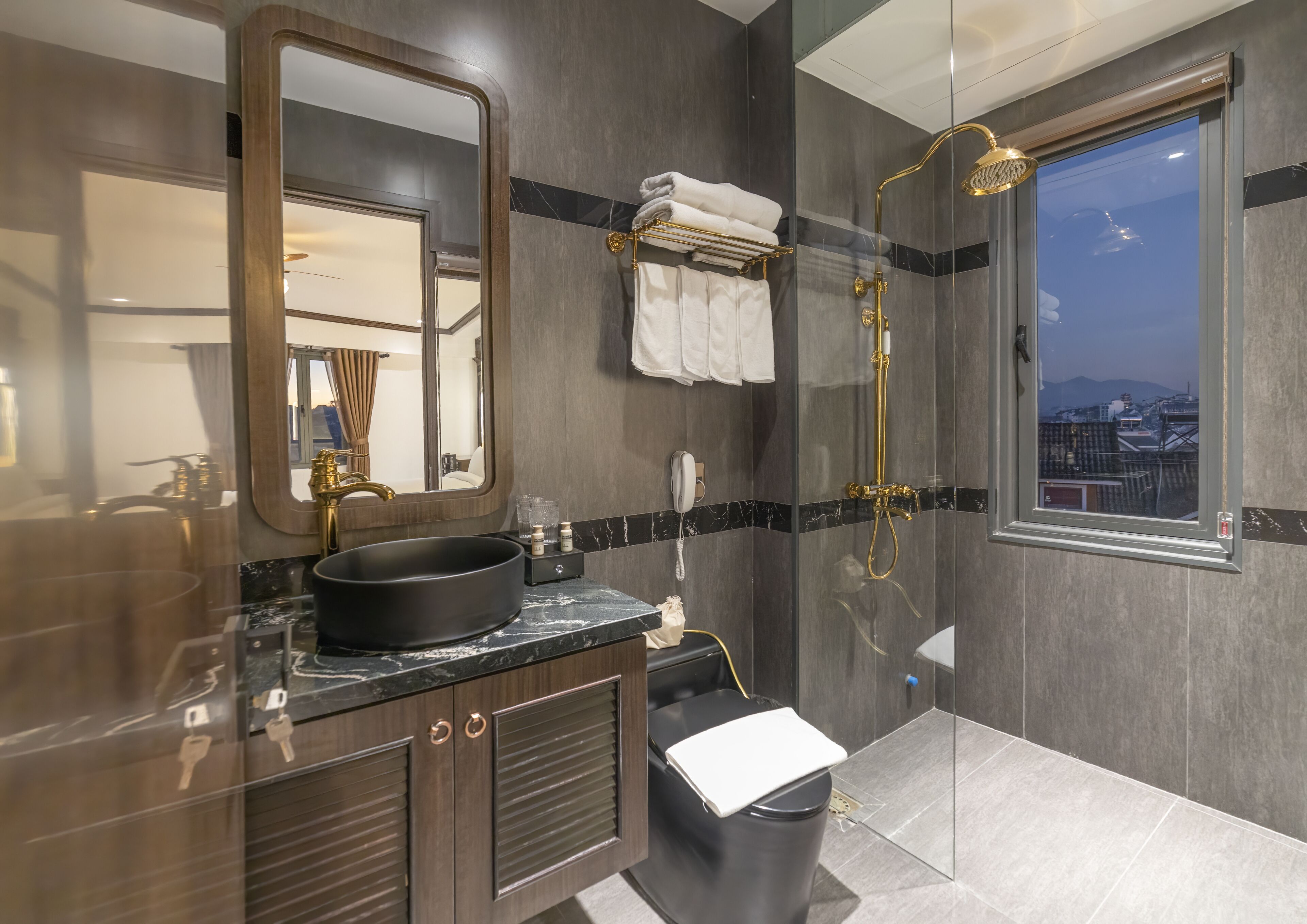 A modern bathroom with a large mirror, a black sink, a gold showerhead, and a view of the city through a window.