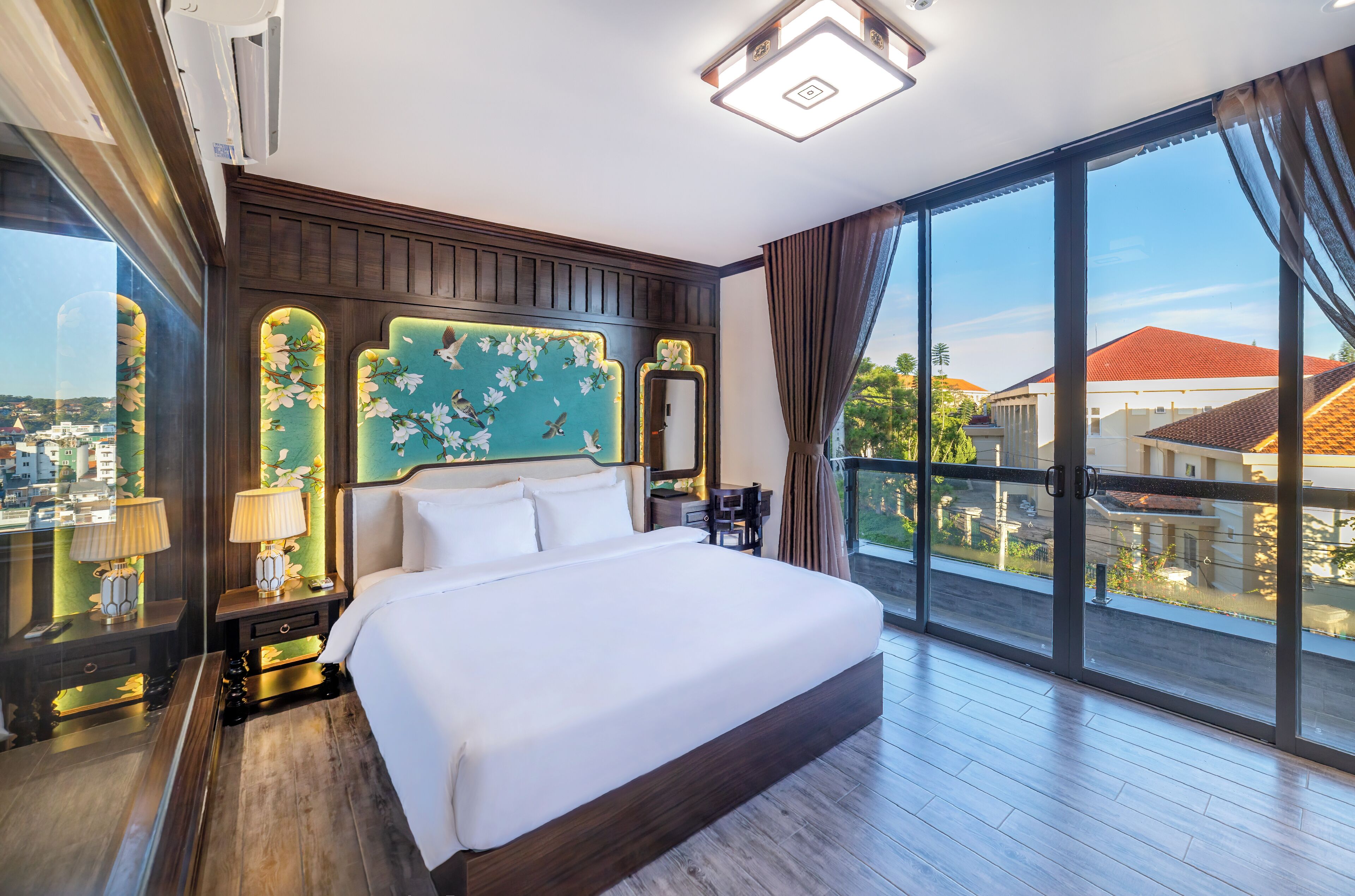 Une chambre d’hôtel avec un grand lit, une table de chevet, un miroir et une vue sur la ville grâce à de grandes fenêtres.