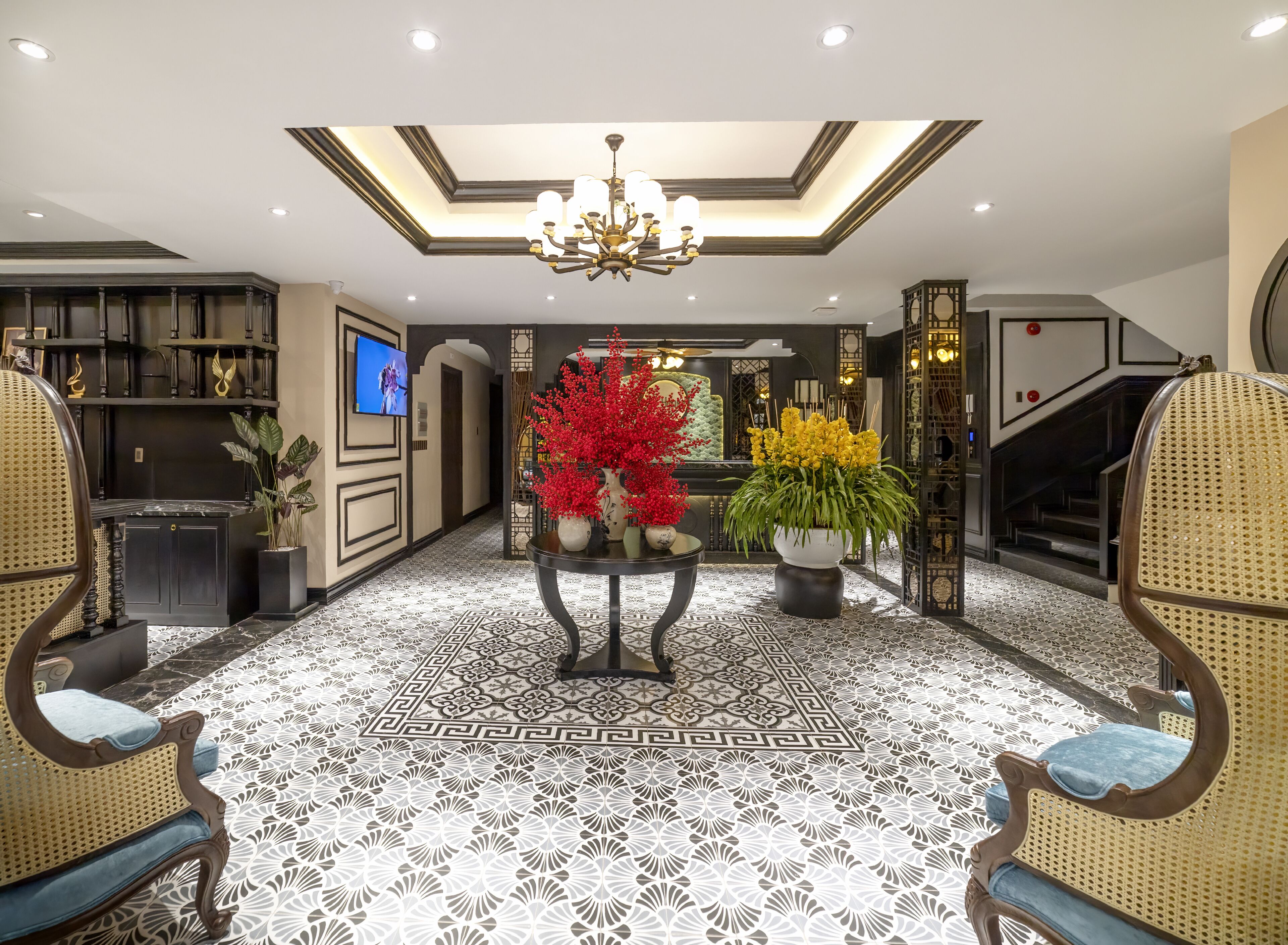 A spacious lobby with a high ceiling, a chandelier, a central table with floral arrangements, and a television mounted on the wall.