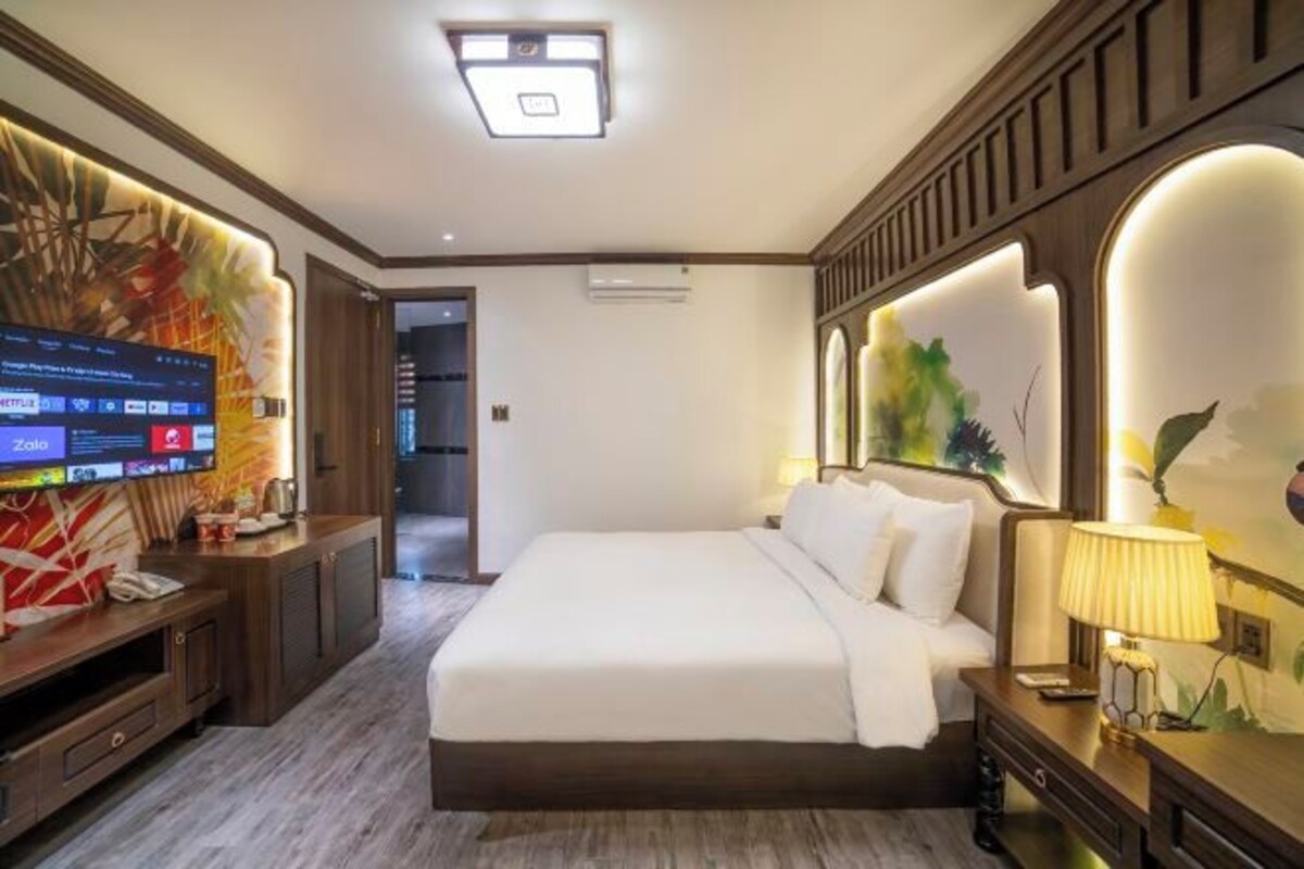 A hotel room with a large bed, a TV on a wooden console, and a wall with a nature-themed design.