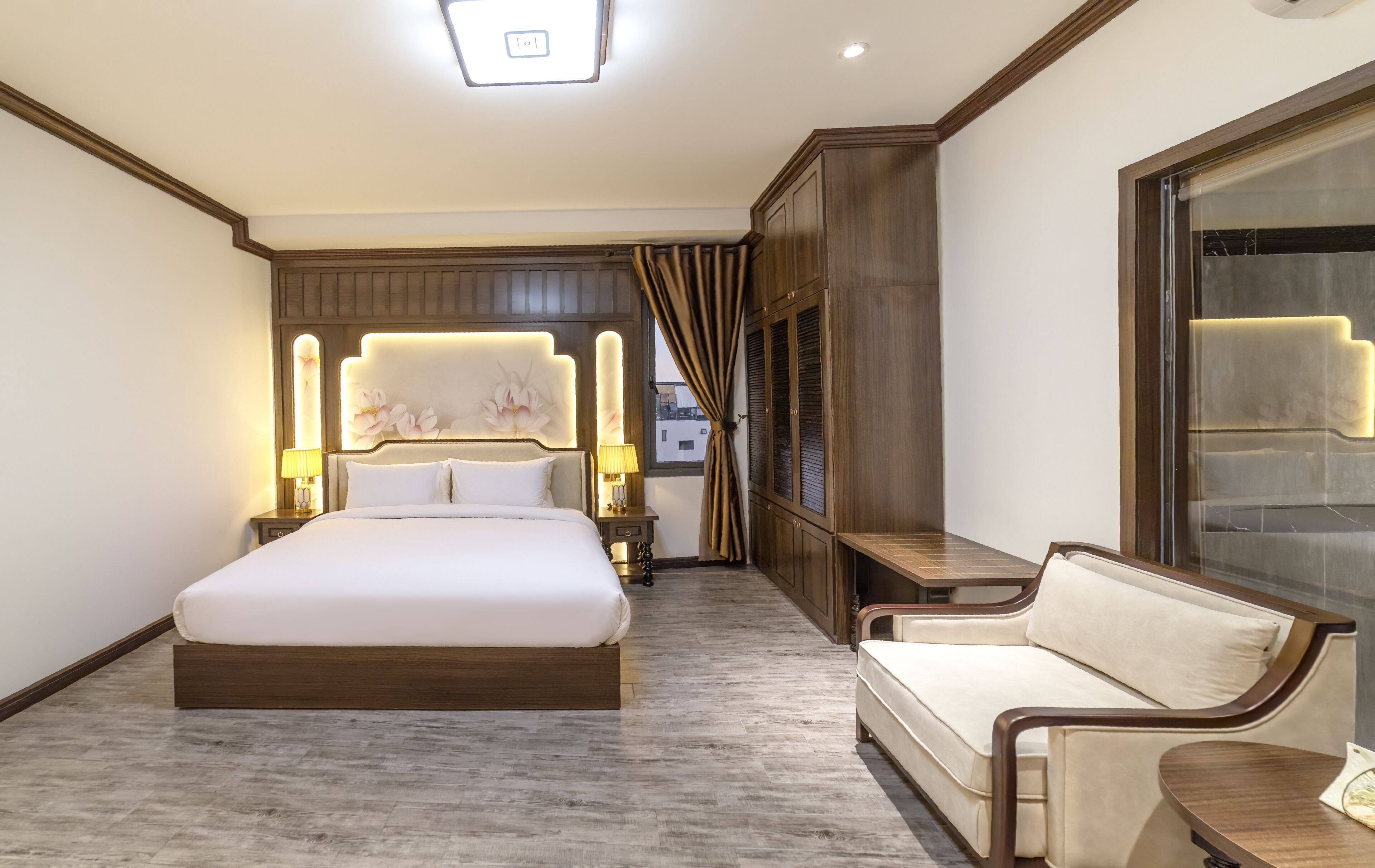 A hotel room with a large bed, a sofa, a nightstand with lamps, a wardrobe, and a large window with curtains.