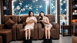Two individuals seated on a brown sofa with black buckets in front of them, in a room with wooden paneling and floral wall art. - Le House Dalat  (Cam Ly - Da Lat)