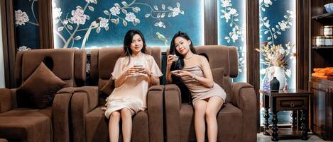 Dos personas sentadas en un sofá marrón, con cubos negros frente a ellas, en una habitación con paneles de madera y decoración floral en las paredes.