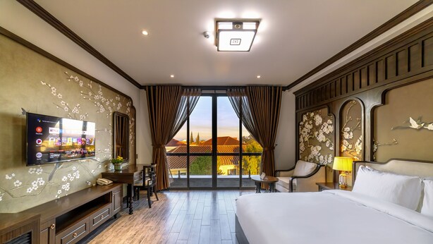 A hotel room with a large bed, a TV on the wall, a desk with a chair, and a view of the outside. - Le House Dalat  (Cam Ly - Da Lat)