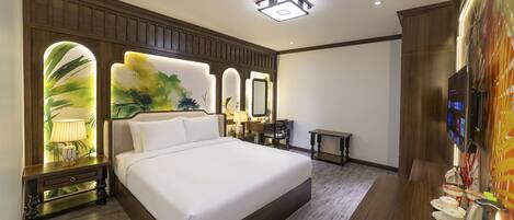 A hotel room with a large bed, a wooden headboard, a nightstand with a lamp, a desk with a chair, and a TV.